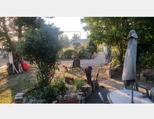 Garten mit verschiedenen Pflanzen und Bäumen, einem zentralen Kiesweg und Sitzgelegenheiten aus Kunststoffstühlen und einem Tisch mit Sonnenschirm. Im Hintergrund ist ein Holzpavillon und eine freie Wiese zu sehen.