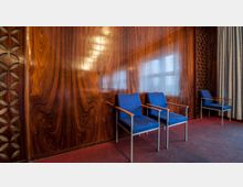 Holzverkleideter Raum mit glänzender Maserung an den Wänden und drei blauen Stühlen mit Armlehnen aus Metall und Holz. Links befinden sich dekorative Holzelemente, rechts ist ein Fenster mit Vorhängen sichtbar.