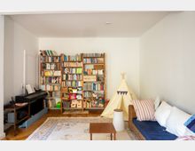 Wohnzimmer mit Holzfußboden, großem Bücherregal an der Rückwand und einem Klavier an der linken Wand. Rechts stehen ein geflochtener Sofa mit Kissen, ein kleiner Couchtisch und ein Tipi-Zelt auf einem gemusterten Teppich.