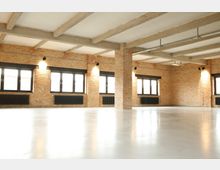 Leerer Raum mit Sichtziegelwänden, großen rechteckigen Fenstern mit schwarzen Rahmen und freiliegenden Betonsäulen und Deckenbalken. Der Boden ist glatt und hell, und unter den Fenstern befinden sich schwarze Heizkörper.