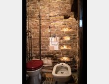 Kleines Badezimmer mit einer freiliegenden Ziegelwand, einem weißen Bidet, einer Toilette mit weinrotem Deckel und sichtbaren Metallrohren. In die Wand sind kleine Nischen mit indirekter Beleuchtung eingelassen.