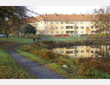 Mehrstöckiges Wohngebäude mit gelber Fassade und roten Ziegeldächern, im Vordergrund ein kleiner Teich mit Spiegelung des Gebäudes sowie ein begrüntes Parkgelände mit Fußweg und vereinzelten Bäumen. Im Hintergrund sind parkende Autos und einige Menschen zu sehen.