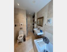 Modernes Badezimmer mit beigefarbenen Wand- und Bodenfliesen. Es ist ausgestattet mit einer Toilette, einem rechteckigen Waschbecken, einer Badewanne und einem großen goldgerahmten Spiegel; verschiedene Pflegeprodukte und Handtücher sind sichtbar.