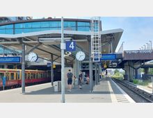 Bahnhof Warschauer Straße in Berlin mit überdachtem Bahnsteig, einer digitalen und analogen Uhr, sowie mehreren Hinweisschildern und Sitzbänken. Ein abgestellter S-Bahn-Zug ist links zu sehen, während rechts die Gleise in die Ferne führen.