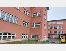 Ein mehrstöckiges Gebäude mit rosafarbenem Putz und weißen rechteckigen Fenstern. Der Hofbereich ist teilweise gepflastert und beinhaltet einen gelben Müllcontainer sowie ein Vordach aus Metall und Wellblech.