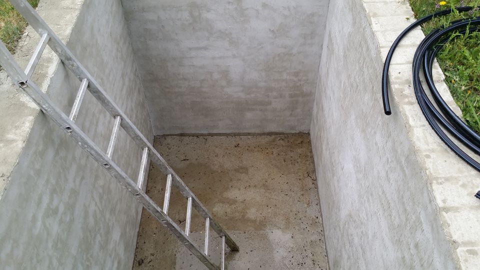 Blick in eine rechteckige, leere Betonbaugrube mit glatten Wänden und Betonboden, in die eine Metallleiter schräg hinabführt. Am oberen Rand ist Gras zu sehen, daneben liegen mehrere aufgerollte schwarze Kunststoffschläuche.