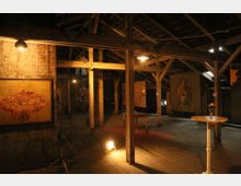 Innenansicht eines rustikalen Dachbodens mit Holzdecken und sichtbaren Balkenkonstruktionen, der als Kunstgalerie genutzt wird. An den Wänden und Balken hängen Gemälde, während einzelne Möbelstücke und ein runder Stehtisch mit Blumenarrangement den Raum ergänzen, der mit warmem Licht beleuchtet ist.