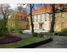 Gelbes Gebäude mit rotem Ziegeldach und hohen Fenstern im klassischen Stil, umgeben von einem kleinen Park mit Bänken, gepflasterten Wegen, Bäumen und Blumenbeeten. Im Vordergrund ist ein Weg aus Steinplatten sichtbar, der entlang gepflegter Grünflächen führt.