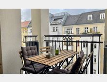 Eine kleine, überdachte Balkonterrasse mit Holztisch und vier gepolsterten Klappstühlen, umgeben von einem Metallgeländer mit dekorativen Elementen. Im Hintergrund Blick auf die Fassaden mehrerer hell gestrichener Altbaugebäude mit Fenstern und Dachflächen.