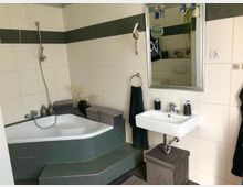 Modernes Badezimmer mit Eckbadewanne, gefliest in Weiß und Dunkelgrün. Neben der Wanne befindet sich ein rechteckiges Waschbecken mit Spiegel und schwarzem Handtuch, umgeben von dekorativen Elementen.