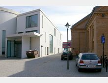 Moderne, weiß verputzte Gebäudefassade mit großen Glasfenstern und Flachdach auf der linken Seite einer engen, gepflasterten Straße; rechts ein Backsteingebäude mit klassischer Architektur, davor geparkte Autos und ein Laternenmast.