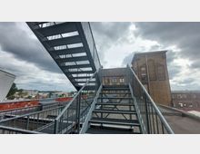 Eine Außentreppe aus Metall führt auf ein Dach mit Blick auf ein historisches Gebäude aus Ziegelstein mit verzierten Rundbogenfenstern. Im Hintergrund sind weitere Gebäude und eine leicht bewölkte Himmelsszenerie zu sehen.