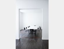 Minimalistischer Besprechungsraum mit einem langen weißen Tisch, umgeben von schwarzen Stühlen. Der Raum hat weiße Wände und einen dunklen Holzboden.