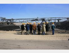 Gruppe von Personen mit gelben Schutzhelmen steht vor einer großen offenen Tagebaugrube, im Hintergrund sind Förderbänder und industrielle Maschinen zu sehen. Der Boden ist sandig, und der Himmel klar.