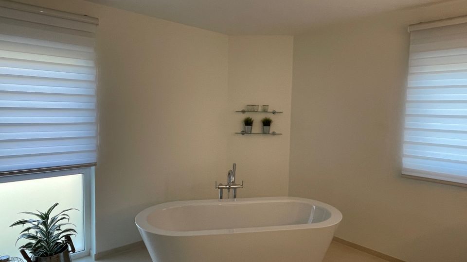 Modernes Badezimmer mit freistehender Badewanne in der Mitte, zwei Fenstern mit halbtransparenten Rollos und einem Regal mit Dekorationen an der Wand. Links unten steht eine Zimmerpflanze in einem silbernen Topf auf dem Boden.