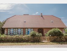 Backsteinhaus mit rotem Ziegeldach, mehreren Dachfenstern und grünen Fensterläden. Vor dem Haus befindet sich ein niedriger Holzzaun mit Pflanzen, und im Vordergrund ist eine gepflasterte Straße erkennbar.