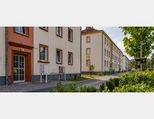 Wohnsiedlung mit mehrstöckigen Gebäuden in heller Putzfassade und orangefarbenen Fensterrahmen sowie einem Gehweg und Grünflächen mit jungen Bäumen und Sträuchern im Vordergrund.