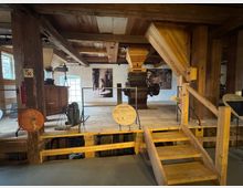 Innenansicht einer historischen Mühle mit Holzbalken, einer Holztreppe und mehreren alten Maschinen. Infotafeln, Schautafeln und ein großes Fenster im Hintergrund ergänzen die rustikale Umgebung.