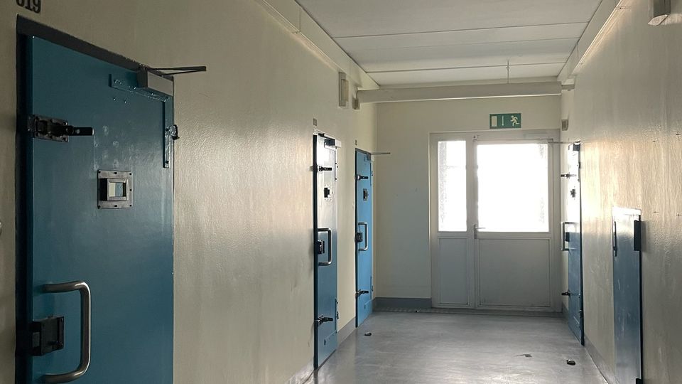 Langer, schmaler Flur in einem Haft- oder Sicherheitsgebäude mit mehreren massiven blauen Stahltüren auf beiden Seiten und glattem grauem Boden. Am Ende des Gangs befindet sich eine helle Doppeltür mit Fluchtweg-Schild darüber.