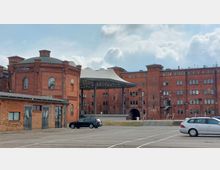 Backsteingebäude eines ehemaligen Industriekomplexes mit runden und eckigen Elementen, teilweise renoviert und mit modernen Erweiterungen wie einem großen, weißen Überdachungselement. Im Vordergrund befindet sich ein Parkplatz mit geparkten Autos, während der Eingang zu einem "Loft Spa" im Hauptgebäude sichtbar ist.