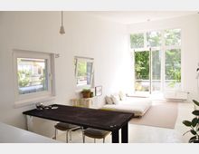 Helles Wohnzimmer mit weißen Wänden, einem dunklen Holztisch und einem niedrigen Sofa mit Kissen auf einem beigefarbenen Teppich. Große Fenster und eine geöffnete Glastür führen zu einem Balkon mit Pflanzen, wodurch viel Tageslicht und Blick auf grüne Bäume hineinkommt.