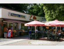 Außenansicht eines kleinen Imbissstandes mit der Aufschrift "Wannsee - Imbiss" und danebenliegendem Eiscafé. Davor stehen Tische mit roten Coca-Cola-Sonnenschirmen und grünen Kunststoffstühlen, umgeben von Bäumen und einer gepflasterten Fläche.