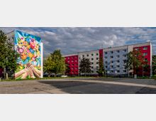 Wohngebiet mit einem farbig gestalteten Plattenbau; die Fassade des linken Gebäudeteils zeigt ein großes Wandgemälde mit ineinander greifenden Händen und bunten Blumen. Im Vordergrund ein gepflasterter Platz, neben dem Bäume und parkende Autos stehen.