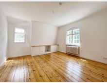 Helles, leerstehendes Zimmer mit weiß gestrichenen Wänden, einem Dielen-Holzboden, zwei Fenstern mit Sprossen und einem Heizkörper darunter. An der Wand befindet sich ein niedriger, eingebauter Schrank mit offenen Regalfächern.