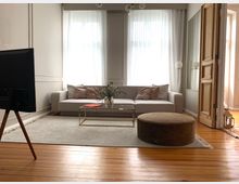 Wohnzimmer mit einem hellgrauen Sofa vor bodentiefen, weißen Vorhängen, einem goldenen Couchtisch, einem braunen runden Hocker und einem Fernseher auf einem Holzständer. Der Raum hat einen Holzboden, eine geöffnete Holztür und dezente Wandverkleidungen.