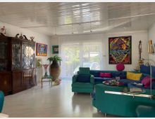 Wohnzimmer mit einer farbenfrohen Einrichtung. Ein großes, türkisfarbenes Ecksofa mit bunten Kissen dominiert den Raum, daneben steht ein Sessel vor einer weißen Wand mit Fenstern, die mit transparenten Vorhängen bedeckt sind. An den Wänden hängen bunte Gemälde, und ein dunkelbrauner Vitrinenschrank steht links. Im Raum befinden sich auch dekorative Elemente wie eine große Vase und eine künstlerisch gestaltete Stuhlskulptur.