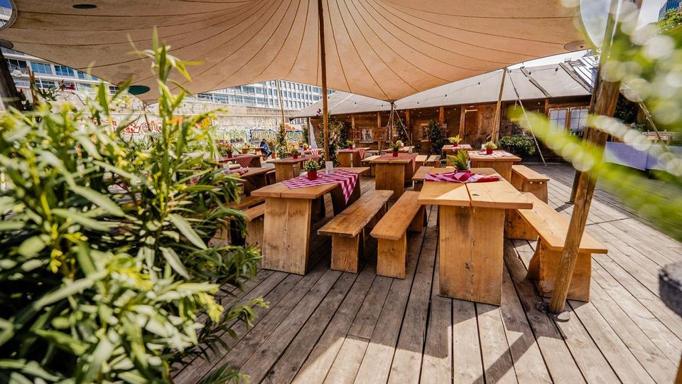 Biergartenähnliche Außenterrasse auf einem Holzdeck mit rustikalen Holztischen und -bänken, teils mit rot-weiß karierten Tischdecken, unter großen Stoff-Sonnensegeln. Im Hintergrund stehen eine Holzbar und Pflanzenkübel, dahinter eine bemalte Wand und moderne Bürogebäude.
