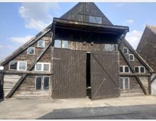 Eine alte Scheune mit dunklen Holzplanken und einem steilen Giebeldach, durchsetzt von vielen kleinen rechteckigen Fenstern. Die zweiflügelige Holztür in der Mitte ist leicht geöffnet, und die Umgebung besteht aus gepflastertem Boden und angrenzenden Gebäuden mit Ziegeldächern.