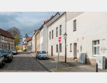 Kopfsteinpflaster-Straße in einer Altstadt mit Reihenhäusern, einigen Fachwerkhäusern und geparkten Autos am Straßenrand. Im Hintergrund ist ein Turm mit spitzem Dach sichtbar, daneben Bäume und weitere Gebäude.