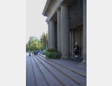 Eingangsbereich eines Gebäudes mit breiter Steintreppe und großen Säulen. Daneben stehen mehrere national gefärbte Flaggen, und im Hintergrund ist ein bemalter Bär sowie grüne Büsche sichtbar.