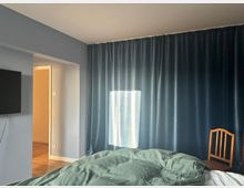 Schlafzimmer mit blauen Vorhängen an der rechten Seite, einem Bett mit grüner Bettdecke im Vordergrund und einem hölzernen Stuhl daneben. Links an der Wand hängt ein Flachbildfernseher, daneben ein Durchgang zu einem hell beleuchteten Flur mit Holzboden.