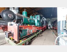 Eine historische Dampflokomotive mit grünem Kessel und rotem Fahrgestell steht in einer Werkstatthalle mit Holzdach. Im Hintergrund sind weitere Lokomotiven und Werkstatteinrichtungen sichtbar.