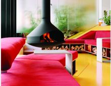 Moderner Wohnraum mit hängendem, schwarzem Kamin in der Mitte, umgeben von pinkfarbenen Polstersofas und gelbem Boden. Im Hintergrund sind Glaswände mit Sichtschutz sowie Pflanzen sichtbar, und unter den Sofas befinden sich Holzstapel als Dekoration.