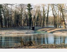 Eine winterliche Parklandschaft mit einem zugefrorenen Teich im Vordergrund und kahlen, teils mit Efeu bewachsenen Bäumen. In der Mitte führt ein kleiner gemauerter Steg über einen schmalen Wasserlauf.