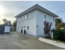 Einfamilienhaus mit hellgrauer Fassade und flachem Dach, umgeben von einem gepflasterten Hof und kleinen Grünflächen mit Sträuchern. Links im Bild befindet sich ein modernes Gartenhaus mit einem Fahrrad darunter.