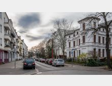 Straßenszene mit mehrstöckigen Wohnhäusern auf der linken Seite und einer historischen weißen Villa mit aufwendigen Verzierungen auf der rechten Seite. Geparkte Autos säumen die Straße, die von kahlen Bäumen und Gehwegen begrenzt wird; es ist bedecktes Wetter.