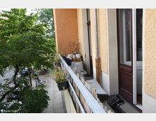 Ein schmaler Balkon eines Wohnhauses mit hellorangefarbener Fassade, ausgestattet mit Blumenkästen, Pflanzentöpfen, Sitzkissen und einem Sichtschutz aus Bambusstäben. Im Hintergrund sind Bäume, Fahrräder sowie Autos entlang eines Gehwegs vor einer Straße erkennbar.
