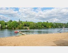 Sandstrand an einem See mit Steg, Badeplattform und einer Wasserrutsche. Im Hintergrund dichter Wald und ein leicht bewölkter Himmel.