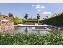 Modern gestalteter Garten mit symmetrischen Wasserbecken, geometrischen Steinterrassen und gepflasterten Wegen. Umgeben von geschnittenen Hecken und Bäumen, unter klarem Himmel mit vereinzelten Wolken.