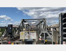 Stählerne Eisenbahnbrücke mit sichtbaren Gleisen und technischen Installationen, umgeben von moderner Stadtbebauung und bewölktem Himmel. Im Vordergrund sind Schilder und ein mit Graffiti bedeckter Kasten erkennbar.