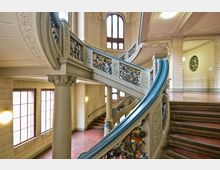 Historisches Treppenhaus mit geschwungener Treppe, aufwendig verzierten Geländern aus Metall und dekorativen floralen Motiven. Große Fenster und hohe Decken prägen den hellen Innenraum, begleitet von verzierten Säulen und ornamentaler Stuckarbeit.