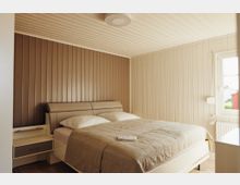 Schlafzimmer mit Holzwänden in Beige- und Brauntönen, einem Doppelbett mit heller Bettwäsche und einem Nachttisch auf der linken Seite. Rechts befindet sich ein Fenster mit Blick nach draußen.