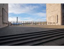 Freifläche mit Treppenstufen zwischen zwei großen Steinmauern, an denen Gedenktafeln angebracht sind; im Hintergrund ein Zaun mit Blick auf ein Stadion und einen hohen, schmalen Turm unter blauem Himmel.