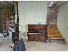 Ein Innenraum mit einem Holzboden und sichtbaren Backsteinwänden. Im Vordergrund steht ein Holzofen mit einem Schornsteinrohr, daneben ein antikes Klavier aus Holz vor einer hell gestrichenen Wand. Rechts führen Holzstufen in den oberen Bereich, links ist ein Regal mit Malerutensilien und eine Leiter zu sehen.