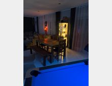 Wohn- und Essbereich mit Holz-Esstisch, Stühlen und Sitzbank auf hellem Fliesenboden, daneben ein graues Sofa und ein beleuchteter Glasvitrinenschrank. Große bodentiefe Fenster mit weißen Gardinen und dunklen Vorhängen säumen die Wand; im Vordergrund ist der Rand eines Billardtischs zu sehen.