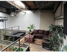 Industriell gestalteter Innenraum mit einer Sitzgruppe aus braunem Sofa, schwarzen Sesseln und Beistelltischen, umgeben von Pflanzen. Der Raum verfügt über eine Betondecke mit Tageslichtfenster und Metallgeländer.